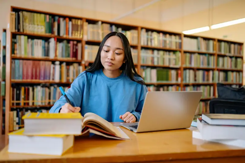 Voksen kvinne som studerer med bøker og bærbar PC i et bibliotek